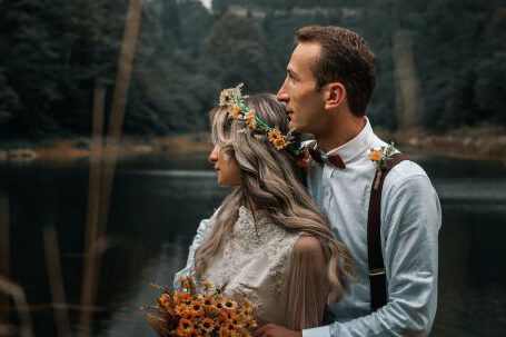 Stylish Wedding Essentials - Young groom gently embracing bride with flower wreath and bouquet while sitting on river shore against woods and looking away